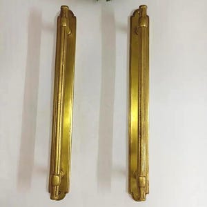May include: Two vintage gold-colored door pulls. Each pull is rectangular with a vertical, ribbed design in the center and decorative ends. The pulls are mounted on a white surface.
