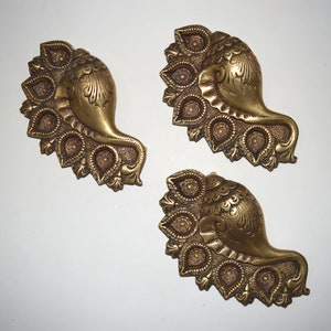 May include: Three gold-coloured metal decorative items shaped like a conch shell with a peacock design. Each shell has a row of small, round indentations along the bottom edge.