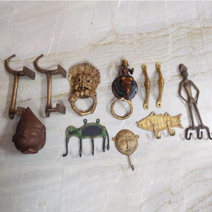 May include: A collection of brass hardware including two L-shaped brackets, a lion head door knocker, a figure-topped ring knocker, two slender handles, a human face mask, an elephant-shaped hook, a fish-shaped hook, and a tall human figure hook.