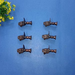 Puede incluir: Seis tiradores de armario en tono bronce con diseño de cabeza de león. Cada tirador presenta una cabeza de león detallada y un mango cilíndrico. Los tiradores están dispuestos sobre una superficie azul, lo que sugiere un tema de ferretería o decoración del hogar.