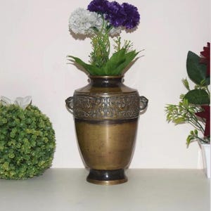 May include: A bronze-colored vase with a decorative band around the top, featuring a floral design. The vase holds a bouquet of artificial flowers, including white and purple carnations. The vase has two handles and a dark base.