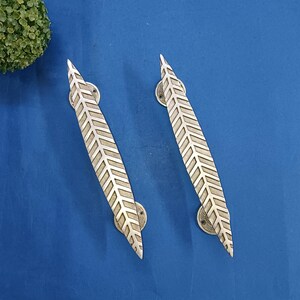 以下が含まれることがあります： 葉のデザインが施された、2つの銀色のドアハンドル。ハンドルは長く細く、平行な線が葉の模様を作り出しています。ハンドルは青い表面に取り付けられています。