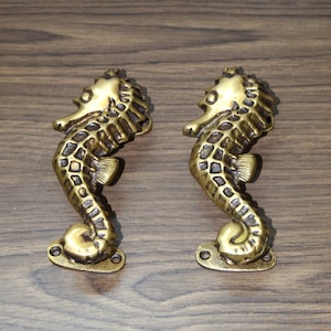May include: Two antique brass seahorse door pulls. Each seahorse is detailed with scales and fins, and mounted on a rectangular base with screw holes. The seahorses are a warm, golden color, and are set against a dark wood grain background.