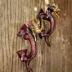以下が含まれることがあります： 2つの装飾的なブロンズ色のドラゴンドアハンドル。 ドラゴンは詳細な鱗を持ち、湾曲したポーズをとっています。 頭は金色の色で、複雑なディテールが施されています。 ハンドルは木製の表面に取り付けられています。