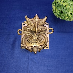 May include: A gold-colored metal door knocker shaped like a mythical creature with a large beak and intricate details. The knocker is mounted on a square plate.