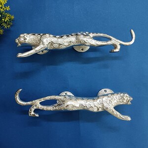 Puede incluir: Dos tiradores de puerta de leopardo plateados. Los tiradores están en pose de carrera, con manchas detalladas y una superficie texturizada. Están montados sobre una superficie azul, con una pequeña planta en la esquina superior izquierda.
