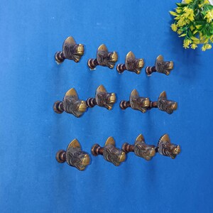 Może przedstawiać: Dziewięć antycznych mosiężnych uchwytów do szafek, każdy ze szczegółowym wzorem głowy zwierzęcia. Uchwyty są zamontowane na niebieskiej powierzchni, prezentując ich skomplikowane rzemiosło i estetykę vintage. Idealne do renowacji mebli.