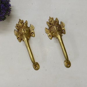 Op de afbeelding: Twee messingkleurige deurgrepen. Elke greep heeft een gedetailleerd olifantenkopontwerp met een kroon en een lange, gebogen handgreep. De handgrepen zijn gemonteerd op een wit oppervlak, wat wijst op een decoratieve of antieke stijl.