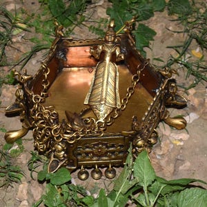 May include: An ornate, gold-colored metal decorative box with a figure in the center. The box has a hexagonal shape with intricate detailing, including chains, small bells, and decorative figures. The box is set on a bed of green foliage.
