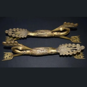 May include: Two antique gold-colored metal tongs with ornate dragon and wave designs. Each tong features a decorative handle with a fan-like end and a three-pronged fork. The tongs are displayed against a dark background.