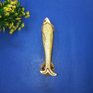 Può includere: Una maniglia per porta a forma di pesce color ottone. La maniglia è dettagliata con squame e pinne ed è montata su una superficie blu. La maniglia è lunga circa 20 cm.