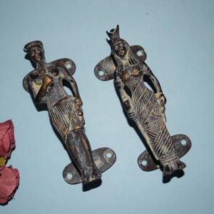 以下が含まれることがあります： 2つのアンティークブロンズ製のドアプルで、それぞれエジプトの女王の様式化された姿が特徴です。彫刻模様と王冠で細部まで表現されています。プルは風化した外観で、取り付け用のプレートが付いています。