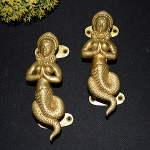 Peut inclure: Deux poignées de porte dorées en forme de sirène. Chaque figurine de sirène a une queue écailleuse détaillée et est en position de prière. Les poignées de porte ont des plaques de montage avec deux trous pour les vis. Le fond est noir.