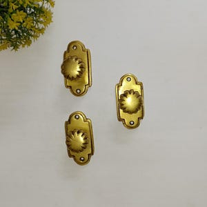 Puede incluir: Tres pomos de puerta dorados con un diseño floral decorativo. Cada pomo está unido a una placa rectangular con un borde festoneado.