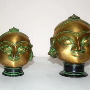 May include: Two antique brass head sculptures with green accents. The sculptures have a stylized face with large eyes, a small nose, and a smiling mouth. The heads sit on a black base. The sculptures are likely decorative items.