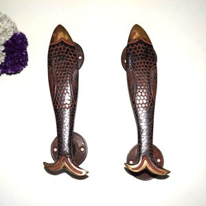 May include: Two decorative door handles in the shape of fish. The handles are made of dark brown metal with a fish scale pattern and gold accents. The handles are mounted on round, bronze-colored plates.