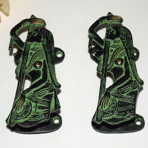 Könnte beinhalten: Zwei antike Türgriffe aus schwarzem und grünem Metall. Jeder Griff zeigt eine detaillierte Silhouette einer Figur, die ein Musikinstrument hält. Die Griffe haben eine Used-Optik und Befestigungslöcher.