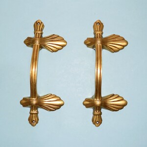 May include: Two gold-toned door handles with a decorative, fan-shaped design. The handles are mounted on a light blue wall.