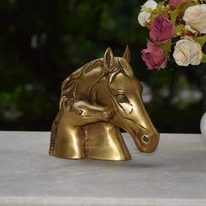 May include: A golden-colored brass sculpture of two horse heads, one larger than the other, positioned side by side. The sculpture is on a white surface, with a blurred green background. A vase of flowers is to the right of the sculpture.
