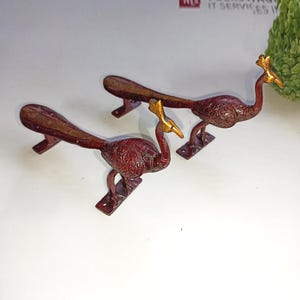 以下が含まれることがあります： 2つの孔雀型の装飾用金属製ハンドル。ハンドルは濃い赤茶色で、頭と尾に金色のアクセントが付いています。ハンドルは長方形のベースに取り付けられています。孔雀の体にはエッチングされたデザインがあります。