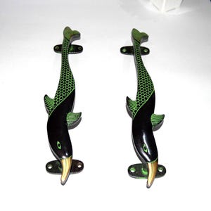May include: Two decorative door handles in the shape of fish. The handles are black with green accents on the fins and tail. The fish's beak is gold. The handles are mounted on a green rectangular base with two holes for screws.