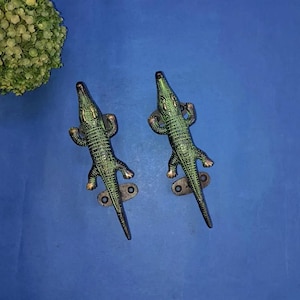 May include: Two green alligator door handles with gold accents. The handles are mounted on a blue background.