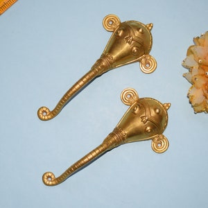 May include: Two ornate, gold-coloured decorative objects with a bulbous head and a long, curved handle. The heads feature intricate detailing and circular accents. The objects are displayed on a light blue surface.