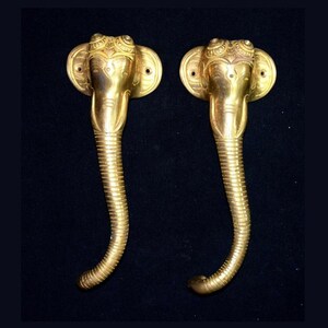 May include: A pair of gold-toned door handles shaped like elephant heads with intricate details. The handles are made of metal and have a traditional Indian design.