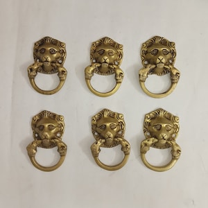 May include: Six gold lion head drawer pulls with a ring handle. The lion heads are detailed with a textured finish.