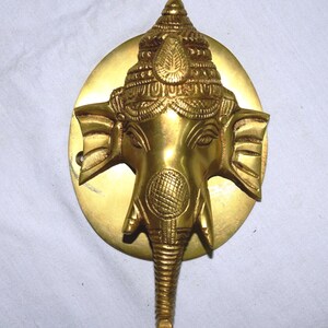 Può includere: Una scultura dorata della testa di Ganesh in ottone. La scultura presenta dettagli intricati, tra cui una corona dettagliata, grandi orecchie e una proboscide. La testa è montata su una base circolare. Il design è ornato e dettagliato.