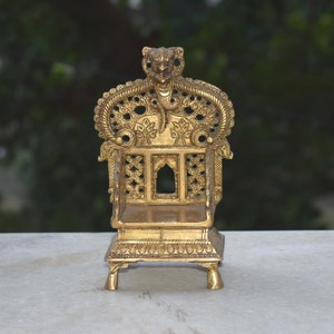Pode incluir: Um pequeno trono de metal dourado adornado com entalhes intrincados e um design detalhado. O trono tem um pequeno espaço aberto nas costas, o que sugere que é destinado a conter um objeto pequeno.