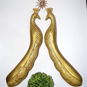 Könnte beinhalten: Zwei goldfarbene, pfauenförmige dekorative Griffe mit aufwendigen Details. Die Griffe sind einander zugewandt, mit einem sternförmigen Ornament darüber. Unten befindet sich ein kleiner grüner Busch.