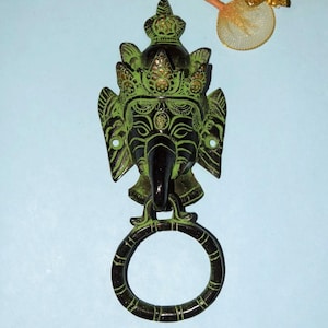 Peut inclure: Un heurtoir de porte en métal vert et noir détaillé, en forme de visage de divinité, avec une trompe d'éléphant et une couronne décorative. Le heurtoir comprend une poignée circulaire en dessous du visage, monté sur une surface bleu clair.