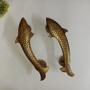 Könnte beinhalten: Zwei Türklinken aus Messing in Fischform. Die Griffe sind gebogen und haben ein detailliertes Schuppenmuster.