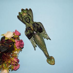 Puede incluir: Una escultura de sirena de color bronce con detalles verdes. La sirena tiene un tocado detallado, alas y una cola escamada. La escultura está sobre un fondo azul claro, con un ramo de rosas en la esquina.