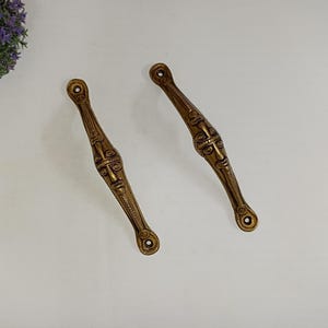 May include: Two antique bronze-colored door handles with a face design. The handles are elongated with a decorative face in the center and holes at each end for mounting. The handles are placed on a white surface.