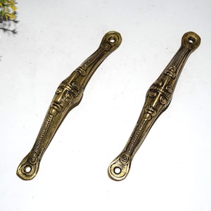 Könnte beinhalten: Zwei antike Türgriffe aus Messing. Jeder Griff weist in der Mitte ein detailliertes Gesichtsdesign mit verzierten Mustern an den Seiten auf. Die Griffe haben eine warme, goldbraune Farbe und sind an jedem Ende mit einem Loch zur Montage versehen.