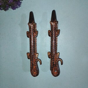Puede incluir: Dos tiradores de puerta decorativos de metal con forma de cocodrilo con acabado negro y marrón. Los tiradores tienen un diseño detallado con detalles dorados.