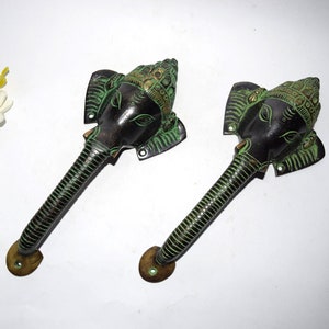 May include: Two antique bronze elephant-head door handles. The handles have a dark bronze finish with green accents, and are detailed with an elephant's face and trunk. The handles are mounted on a white surface.