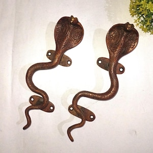 May include: A pair of antique bronze-coloured door handles, each crafted in the shape of a cobra. The handles feature detailed cobra heads and curved bodies, with mounting plates. The metal handles have an aged appearance.
