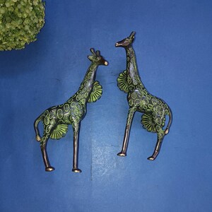 以下が含まれることがあります： 緑と茶色のペイントが施された真鍮製のキリンの置物2つ。キリンは青い表面に立っており、互いに向き合っている。