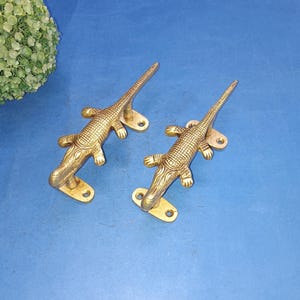 May include: Two gold-toned alligator-shaped door handles. The handles are detailed with textured scales and have a polished finish.