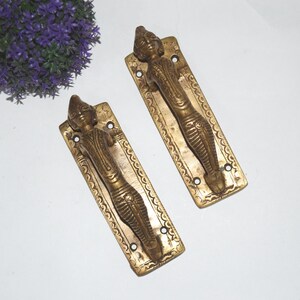 May include: Two antique brass door handles with intricate designs featuring a figure in a reclining pose. The handles are rectangular and have a decorative border.