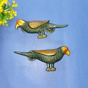 Puede incluir: Dos tiradores decorativos con forma de loro. Los tiradores son de metal con cuerpo verde, alas doradas y pico amarillo. Están diseñados para montarse en un armario o cajón, añadiendo un toque decorativo.