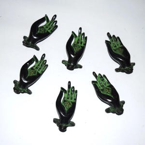 May include: Six small, black hand sculptures with green accents. Each hand is in a different gesture, with intricate details on the palms and fingers. The hands are arranged in a circular pattern against a white background.