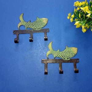 May include: Two metal wall hooks with a fish design. The fish are green and gold, and the hooks are black. Each hook has three hooks for hanging items. The hooks are mounted on a blue surface.