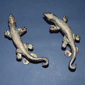 May include: Two bronze-colored lizard-shaped door handles. Each handle features a detailed scale pattern and is mounted on a circular base with screw holes. The lizards are positioned as if crawling, with their tails curled.