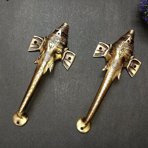 May include: Two gold-toned door handles shaped like the head of an elephant god with intricate details. The handles are made of metal and have a traditional Indian design.