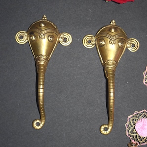 May include: Two brass-colored elephant-shaped door handles. Each handle features a detailed design of an elephant head with large ears and a trunk that curves into a spiral. The handles are likely for decorative purposes.