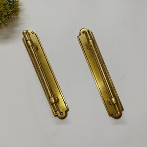 May include: Two gold-toned metal door pulls with a rectangular shape and a decorative design. The pulls have a smooth, polished finish.